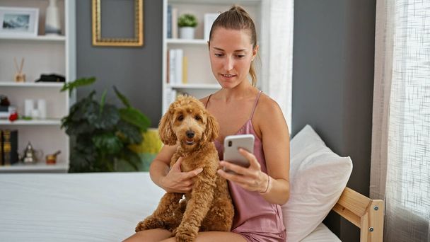 寝室でベッドに座ってスマートフォンを使用して犬を飼っている若い白人女性 - 写真・画像