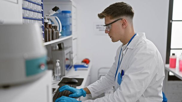 Houkutteleva nuori valkoihoinen miespuolinen tiedemies, joka on täysin uppoutunut tietokoneen kirjoittamiseen koeputkien ja mikroskooppien keskellä, sisätiloissa tehtävä laboratoriotyö - Valokuva, kuva