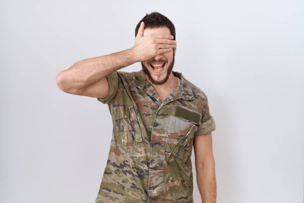 Young hispanic man wearing camouflage army uniform smiling and laughing with hand on face covering eyes for surprise. blind concept.  - Zdjęcie, obraz