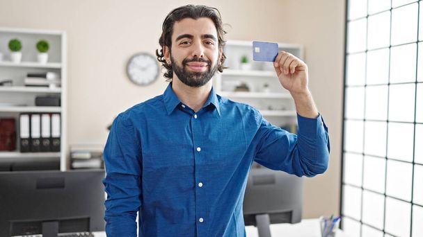 Jeune homme d'affaires hispanique tenant une carte de crédit souriant au bureau - Photo, image