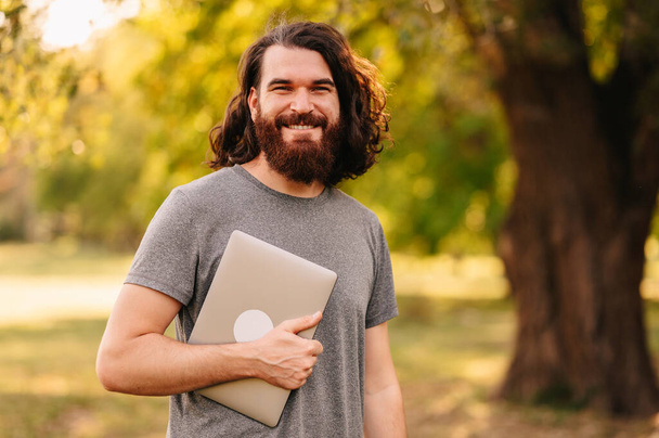 joyful young bearded hipster man holding laptop in park and looking at camera. - Photo, Image