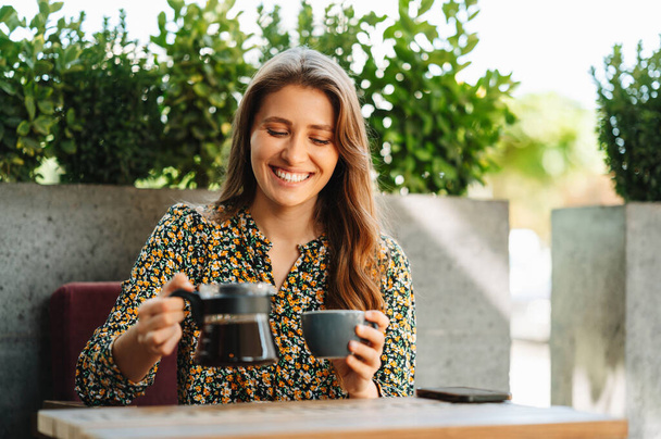 Charming young woman pouring her morning coffee in cafe outdoors. - Photo, Image