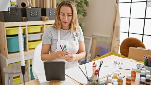 Attraktive junge blonde Künstlerin blendet, während sie mit einem Touchpad eine Online-Zeichenstunde in ihrem Kunstatelier unterrichtet - Foto, Bild