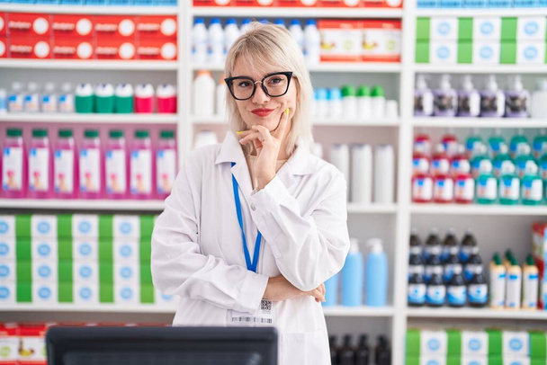 Giovane donna caucasica che lavora in farmacia farmacia guardando fiduciosa la fotocamera sorridente con le braccia incrociate e la mano sollevata sul mento. pensare positivo.  - Foto, immagini