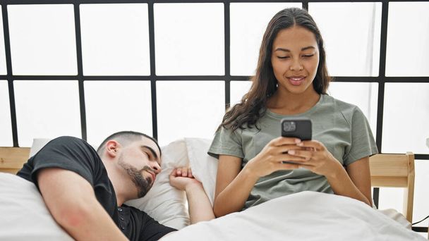 Krásný pár útulný v posteli, přítelkyně na smartphone, zatímco spí přítel osvětluje pokoj, vychutnává jejich klidné vnitřní ráno spolu - Fotografie, Obrázek