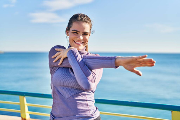 Jovem bela mulher hispânica vestindo sportswear esticando o braço à beira-mar - Foto, Imagem