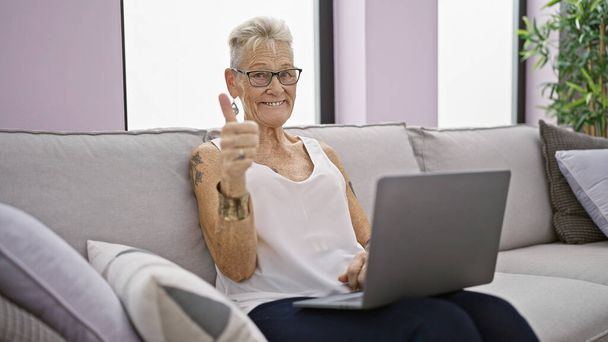 Veselá, šedovlasá starší žena sedící doma na pohovce, nechávajíc dobré vibrace proudit při použití notebooku a signálního gesta palcem nahoru, obklopená šťastnou techno atmosférou - Fotografie, Obrázek