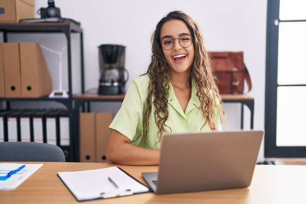 Mladá hispánka pracující v kanceláři v brýlích mrkající na kameru se sexy výrazem, veselou a šťastnou tváří.  - Fotografie, Obrázek