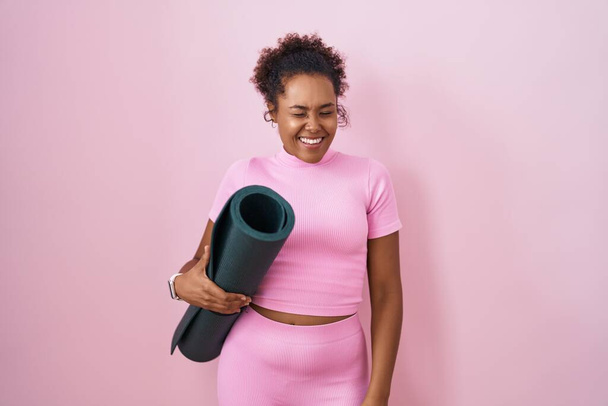 Junge hispanische Frau mit lockigem Haar hält Yogamatte über rosa Hintergrund und zwinkert in die Kamera mit sexy Ausdruck, fröhliches und glückliches Gesicht.  - Foto, Bild