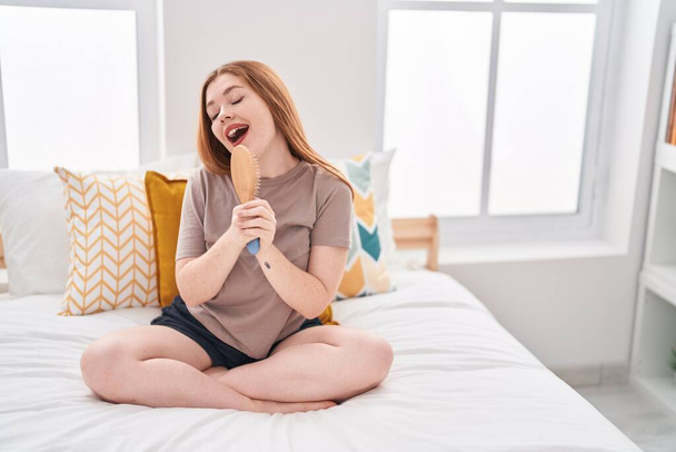 Mujer pelirroja joven cantando canción usando cepillo como micrófono en el dormitorio - Foto, Imagen