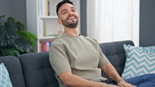 Νεαρός Ισπανός άνδρας χαμογελά σίγουρος κάθεται στον καναπέ στο σπίτι - Φωτογραφία, εικόνα