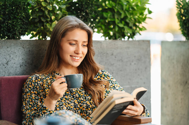 Zamknij zdjęcie młodej uroczej kobiety siedzącej w kawiarni i czytającej książkę i trzymającej filiżankę dobrej kawy. - Zdjęcie, obraz