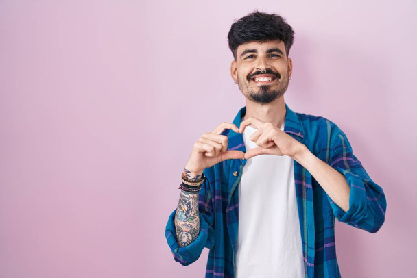 ピンクのバックグラウンドの上に立っているひげを持つ若いヒスパニック男性は,ハートシンボルを手で形作るのが大好きです. ロマンチックなコンセプト.  - 写真・画像