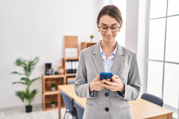 Mujer joven caucásica trabajadora de negocios sonriendo confiada usando teléfono inteligente en la oficina - Foto, Imagen
