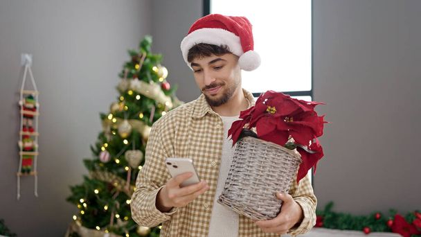 Young arab man using smartphone holding plant standing by christmas tree at home - Photo, Image