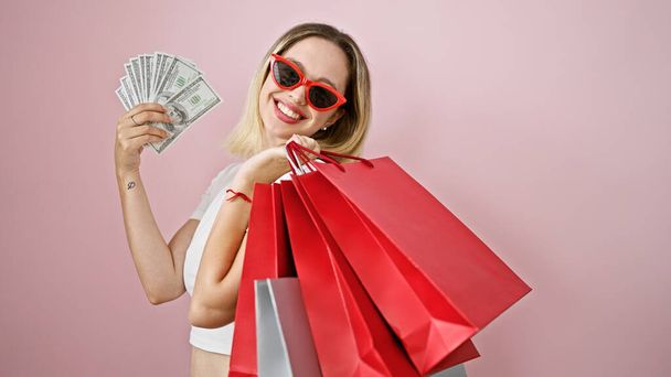 Junge blonde Kundin mit Einkaufstaschen und Dollars vor isoliertem rosa Hintergrund - Foto, Bild