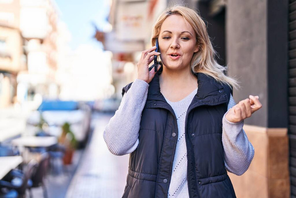 Νεαρή ξανθιά γυναίκα χαμογελά αυτοπεποίθηση μιλώντας στο smartphone στο δρόμο - Φωτογραφία, εικόνα