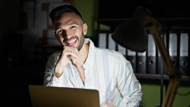 Młody latynoski pracownik biznesowy korzystający z laptopa uśmiechnięty w biurze - Zdjęcie, obraz