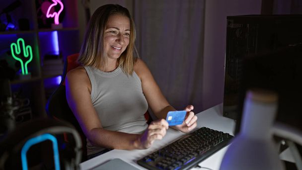 Levendige, jonge blonde streamer meisje live-streaming gaming sessie van gezellige thuiskantoor, glimlachen als ze gebruikt credit card op haar computer in de hitte van de nacht - Foto, afbeelding