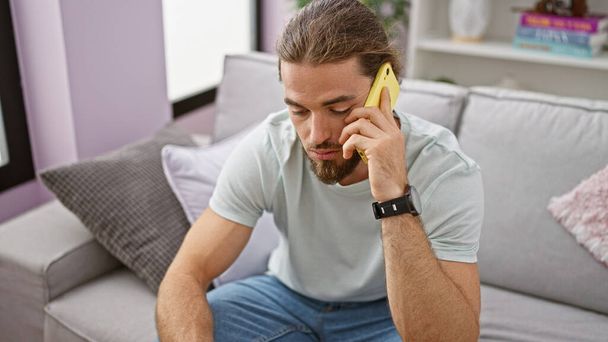 Giovane uomo ispanico che parla al telefono seduto sul divano di casa - Foto, immagini