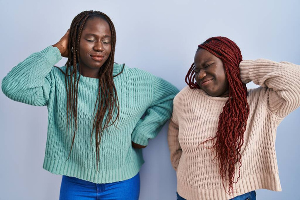 Mavi arka planda dikilen iki Afrikalı kadın boyun ağrısı çekiyor, eliyle boynuna dokunuyor, kas ağrısı çekiyor.  - Fotoğraf, Görsel