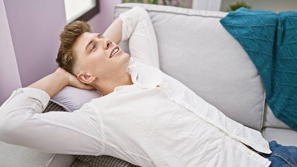 Handsome young caucasian man relaxing in the cozy living room, confidently lying on the sofa with a joyful smile. his hands resting on his head, enjoying the sunlight at his comfortable home. - Photo, Image