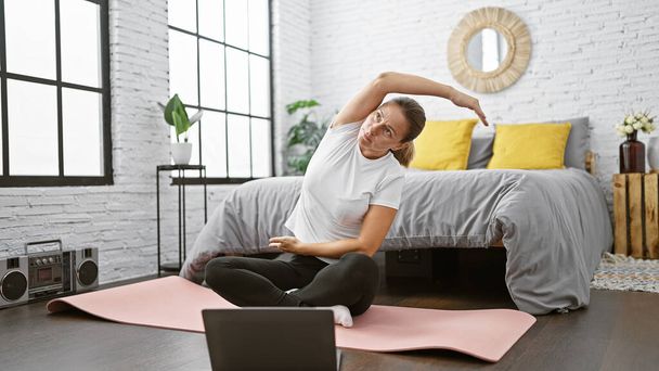 Mujer joven atractiva sonriendo cálidamente mientras posa en una sesión de estiramiento matutino en el suelo de su dormitorio, participando en una vigorizante clase de ejercicios en línea con su computadora portátil - Foto, imagen