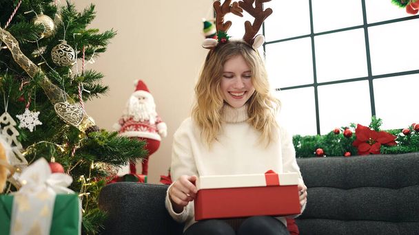 Mladá blondýna žena rozbalování dárek sedí na pohovce u vánočního stromku doma - Fotografie, Obrázek
