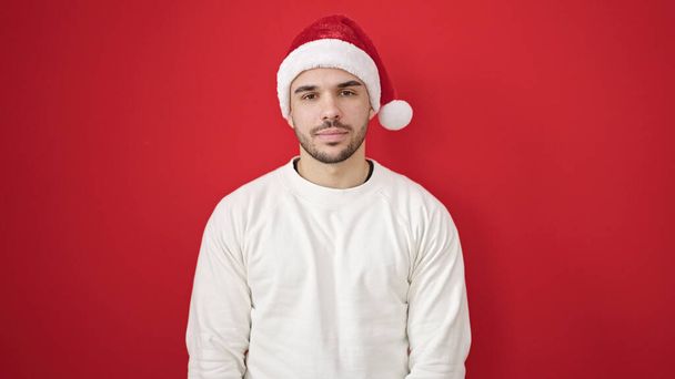 Joven hombre hispano sonriendo confiado usando sombrero de Navidad sobre fondo rojo aislado - Foto, Imagen
