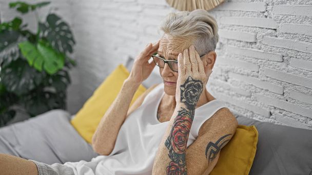 Gestresste grauhaarige Seniorin, die unter Kopfschmerzen leidet, sitzt auf dem Bett in ihrem Schlafzimmer und drückt Besorgnis und Erschöpfung aus - Foto, Bild