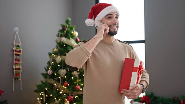 スマートフォンで話す若いアラブの男は,自宅でクリスマスツリーで立っているギフトを保持しています - 写真・画像