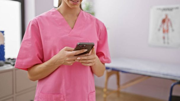 Χαμογελαστή θεραπεύτρια γυναίκα χρησιμοποιεί με αυτοπεποίθηση smartphone κατά τη διάρκεια της συνεδρίας αποκατάστασης, παρέχοντας πρακτική φυσιοθεραπεία θεραπεία στο ζεστό εσωτερικό της κλινικής - Φωτογραφία, εικόνα