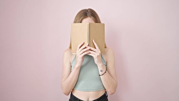 Junge blonde Frau bedeckt Gesicht mit Buch über isoliertem rosa Hintergrund - Foto, Bild