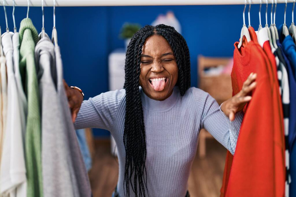 Αφροαμερικανή γυναίκα που ψάχνει ρούχα στο ράφι με ρούχα να βγάζει τη γλώσσα της χαρούμενη με αστεία έκφραση.  - Φωτογραφία, εικόνα