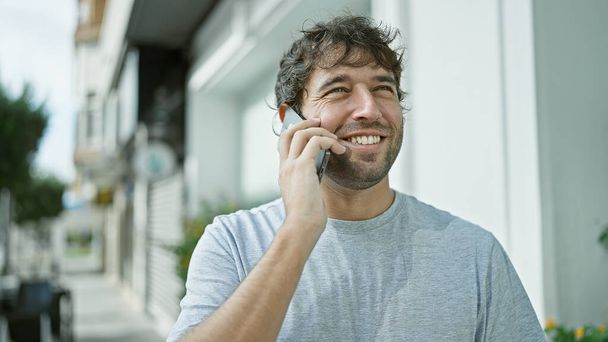 Fröhlicher junger Mann telefoniert, blinkt sein Lächeln auf einem lässigen Straßenbummel - Foto, Bild
