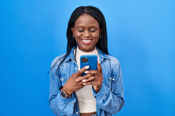 Örgülü Afrikalı kadın akıllı telefon kullanarak mesaj yazıyor. Pozitif ve mutlu görünüyor. Dişlerini gösteren bir gülümsemeyle ayakta duruyor ve gülümsüyor.  - Fotoğraf, Görsel