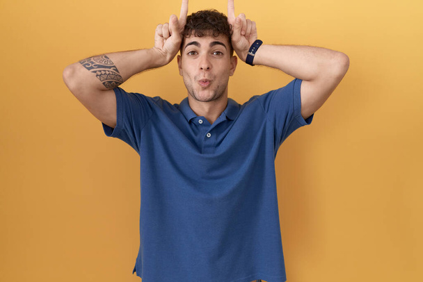 Young hispanic man standing over yellow background doing funny gesture with finger over head as bull horns  - Фото, изображение