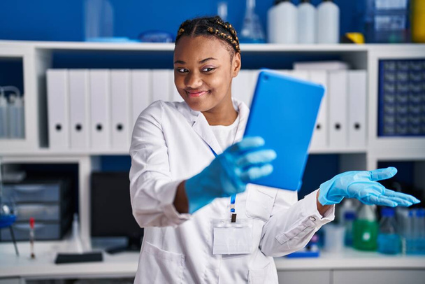 Afro-Amerikaanse vrouw met vlechten werken op wetenschapper laboratorium doen video call met tablet vieren prestatie met een vrolijke glimlach en winnaar uitdrukking met opgeheven hand  - Foto, afbeelding