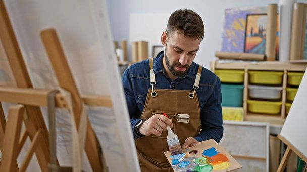 Ontspannen jonge Spaanse man, een portret van een knappe bebaarde kunstenaar, gefocust en geconcentreerd op tekenen met penselen in kunststudio, perfectioneren canvas meesterwerk - Foto, afbeelding