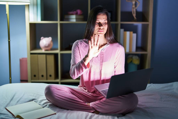 Mujer hispana joven usando computadora portátil en la cama haciendo dejar de cantar con la palma de la mano. expresión de advertencia con gesto negativo y grave en la cara.  - Foto, imagen