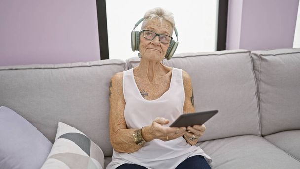 Femme âgée aux cheveux gris confortable, relaxante, assise sur un canapé du salon, captivée par la musique qu'elle écoute à la maison, entourée de technologie - Photo, image