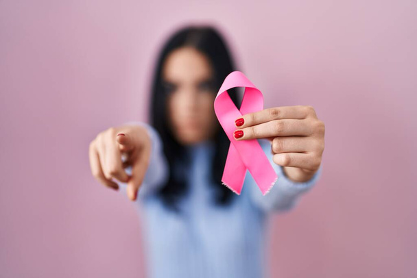 Spanyol nő kezében rózsaszín rák szalag mutató ujjal a kamera és az Ön számára, magabiztos gesztus néz komoly  - Fotó, kép