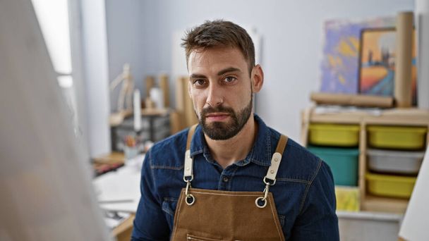 Zaostřený mladý hispánský umělec sedící uvnitř uměleckého studia, pohledný vousatý muž zapojený do vážné malby štětcem na plátno - Fotografie, Obrázek