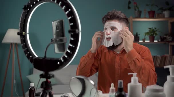 Férfi szépségblogger. Vlogger alkalmazása szépség lap maszk, miközben rögzíti a videót, és így tippeket a szépség blog. Fiatal szakállas férfi teszteli az új terméket. Kozmetikai lepedő maszk. Fénymásolási hely - Felvétel, videó