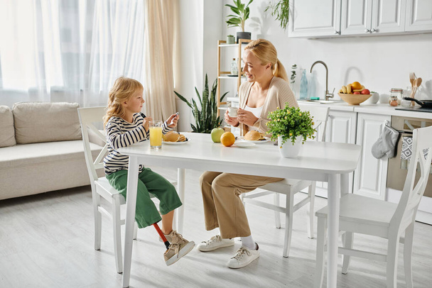 Protez bacaklı anaokulu kızı modern mutfakta mutlu sarışın annesiyle kahvaltı yapıyor. - Fotoğraf, Görsel