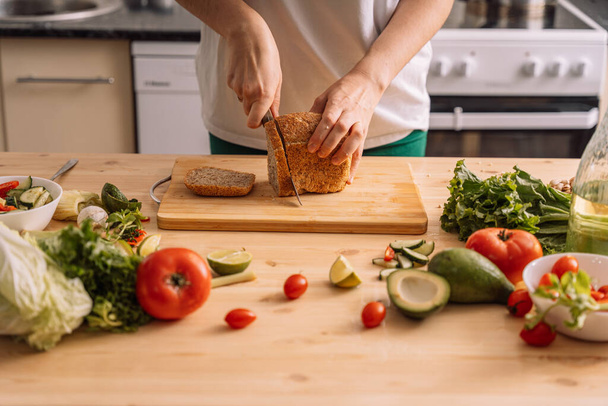 Lady szeletelő kenyeret egy vegetáriánus szendvics egy fa asztalon között friss zöldségek. - Fotó, kép