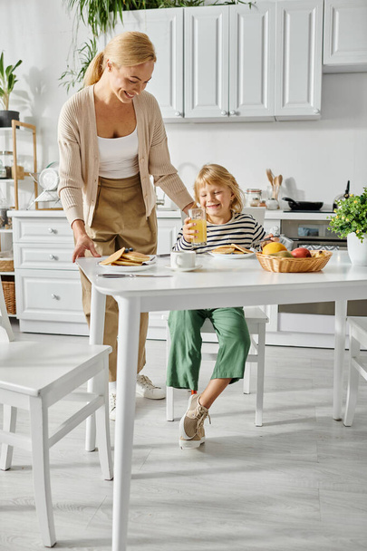 happy woman serving pancakes to daughter with prosthetic leg during breakfast in kitchen, inclusion - Photo, Image