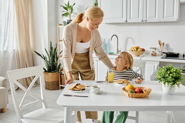 mãe feliz olhando para a filha bonito com perna protética durante o café da manhã na cozinha, inclusão - Foto, Imagem