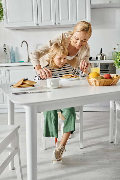 ευτυχισμένη γυναίκα στέκεται πίσω από χαριτωμένο κόρη με προσθετικό πόδι κατά τη διάρκεια του πρωινού στην κουζίνα, ένταξη - Φωτογραφία, εικόνα