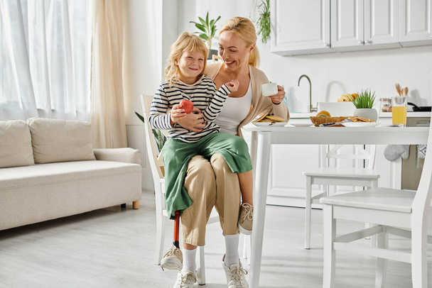 menina pré-escolar com perna protética sentado em voltas de mãe feliz durante o café da manhã na cozinha - Foto, Imagem